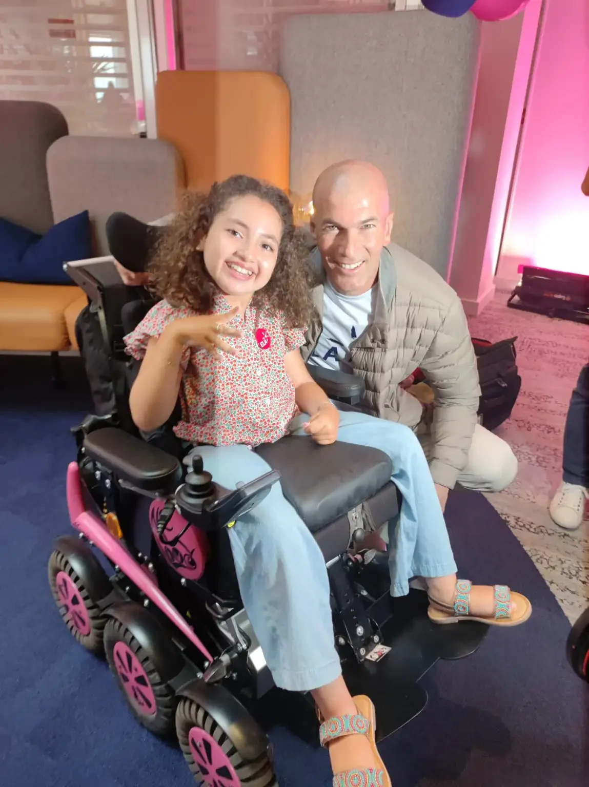 Une jeune fille dans son fauteuil et un adulte sourient ensemble à l’appareil photo.