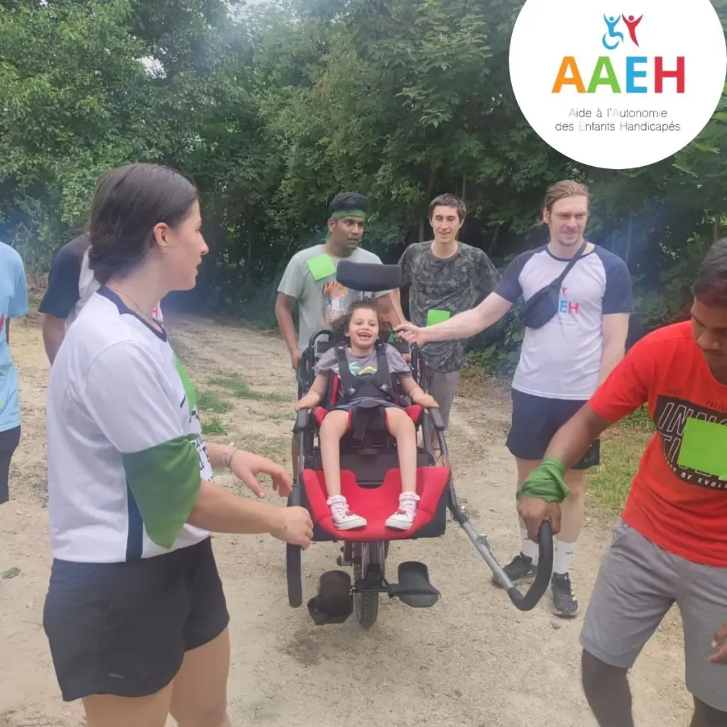 Un groupe accompagne une jeune participante dans un fauteuil de randonnée sur un chemin bordé d’arbres.